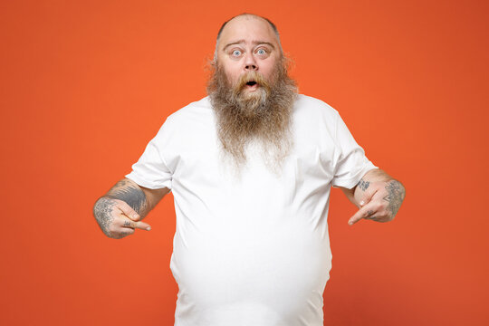 Fat surprised pudge obese chubby overweight tattooed blue-eyed bearded man has big belly in white t-shirt point index finger down on workpase mock up isolated on red orange background studio portrait.