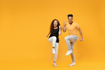 Fototapeta na wymiar Full length young couple together two friends family smiling african man woman in black tshirt do winner gesture clench fist with raised up leg celebrate isolated on yellow background studio portrait