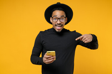 Young surprised shocked excited happy american african man 20s in stylish black shirt hat eyeglasses point index finger on mobile cell phone isolated on yellow orange color background studio portrait