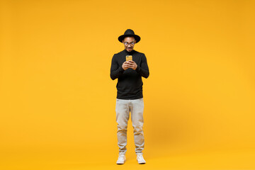 Full length young friendly smiling happy american african man 20s in stylish black shirt hat eyeglasses using mobile cell phone chat in social network isolated on yellow background studio portrait