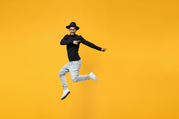 Full length young fun african man 20s in stylish black hat shirt eyeglasses jump high point index finger aside on copy space area mock up isolated on yellow orange color background studio portrait