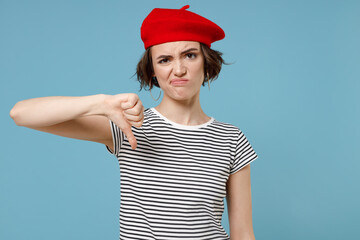 Young displeased dissatisfied european woman 20s with short hairdo wear french beret red hat striped t-shirt show thumb down dislike gesture isolated on pastel blue color background studio portrait.