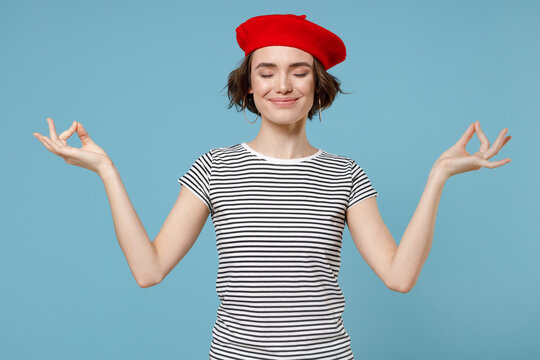 Young spiritual woman 20s with short hairdo in french beret red hat striped tshirt hold spread hands in yoga gesture relax meditate try to calm down isolated on pastel blue background studio portrait