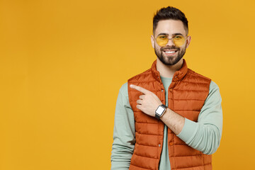 Young smiling happy fun man 20s wear orange vest mint sweatshirt glasses point index finger forefinger aside on copy space area mock up workspace isolated on yellow color background studio portrait