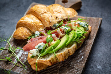 Croissant sandwich with prosciutto ham or jamon, blue cheese, avocado, microgin on blue background. Delicious snack