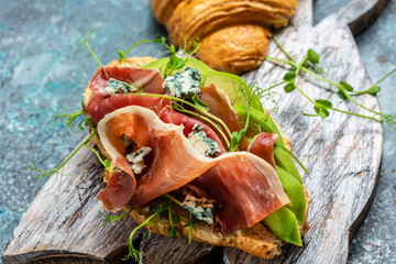 Croissant sandwich with prosciutto ham or jamon, blue cheese, avocado, microgin on blue background....