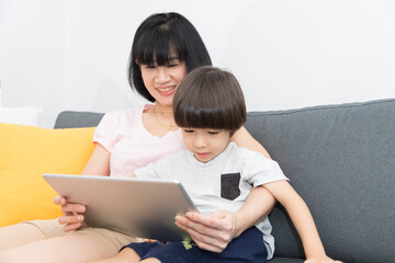 Fototapeta premium Cheerful Asian mother with little son sitting on sofa and playing video game on tablet while spending time together at home.