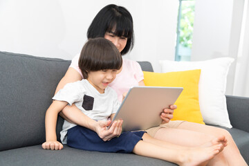 Cheerful Asian mother with little son sitting on sofa and playing video game on tablet while spending time together at home.