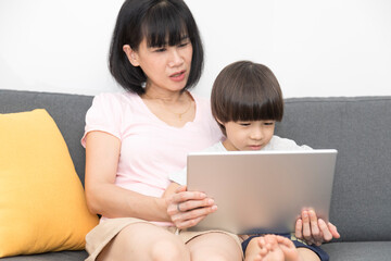 Cheerful Asian mother with little son sitting on sofa and playing video game on tablet while spending time together at home.