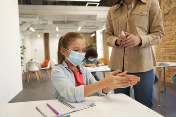 Attractive little school girl wearing protective mask cleaning her hands. Female teacher using an alcohol spray to disinfect student hands in classroom