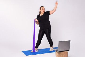 Home activity and training, online fitness class. Overweight blonde woman doing stretching exercises, violet resistance band watching video guide, blue fitness mat, using laptop, white background