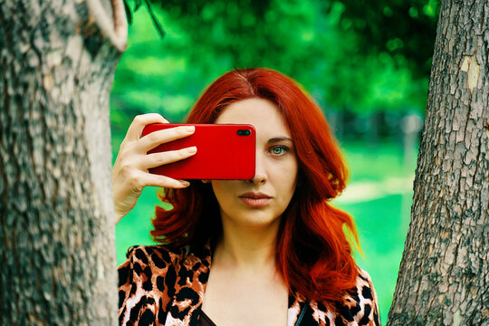 Girl in the Park. Red-haired woman takes a picture on her phone between the trees. Summer walk in the park. An example of excellent camera quality. Differences between phone camera and human eye.