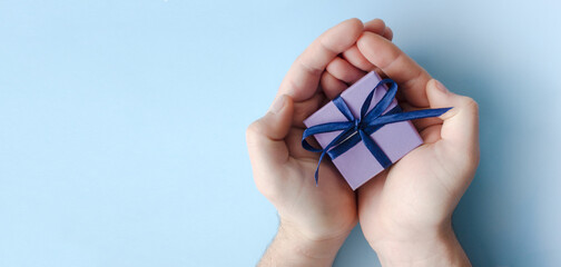 Purple gift box with blue bow in male hands on light blue background. Top view. Copy space.