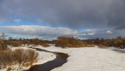Spring landscape with a river and melting snow on the river bank. Dramatic sky. Trees and bushes on the horizon. Early spring.
