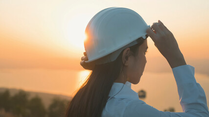 a woman engineer is putting a protective helmet on her head at sunset...