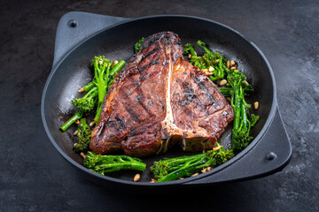 Modern style traditional barbecue dry aged wagyu porterhouse beef steak bistecca alla Fiorentina with baby broccoli and pine nuts served as close-up in a cast-iron design skillet