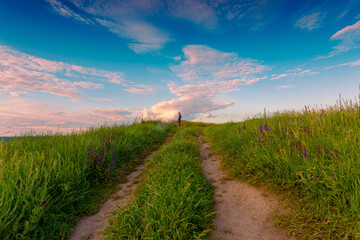 Young man, as small detail, standing on a spring grass hill with the rural road, under the beautiful evening sky with clouds. He looking away and enjoying the moment.