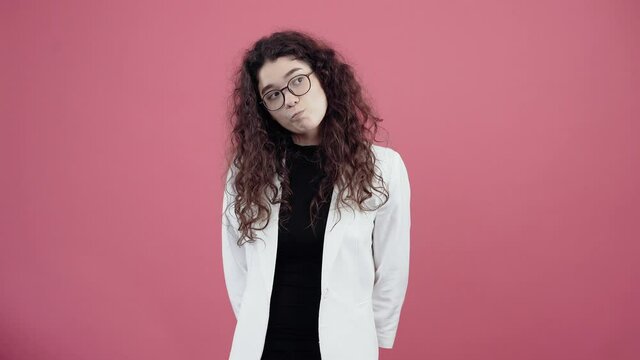 The intelligent young woman with curly hair is thinking of new ideas and strategies for solving a problem. Young hipster in white jacket and black shirt, with glasses posing isolated on pink