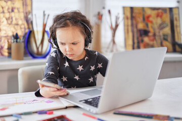 A small kid girl in handsfree headphones sitting at a table using a laptop computer and smartphone....