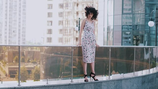 A slender girl with curly hair on a bridge near tall buildings. The wind blows through your hair