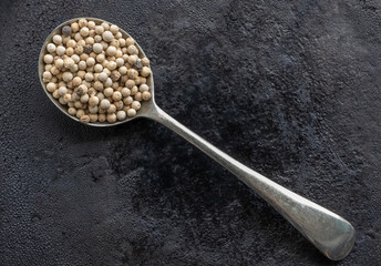 White peppercorns in a deep cupronickel vintage spoon on a black textured background. Spices and herbs.