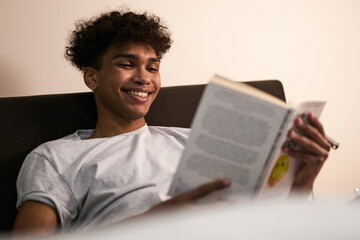 Amazing story. Portrait of cheerful brunette guy lying in the bed at home, smiling while reading interesting book at late evening