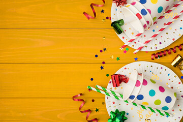 Obraz premium Overhead photo of cups drink with party tubules whistle multicolored confetti bow and plates isolated on the yellow wooden background with empty space