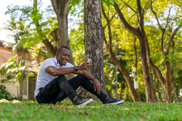 Naklejka na ściany i meble Portrait of a happy African American man using a cellular mobile phone in the garden park. Concept of 5G wireless technology, High-speed Internet, Global network telecommunication, Smart communication