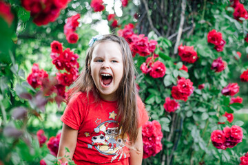 close-up portrait of a screaming, overjoyed girl with her mouth wide open in a red T-shirt near a rose bush in a park or garden.