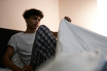 Relaxation. Young guy wearing pajamas going to bed, covering himself with a duvet in the bedroom - obrazy, fototapety, plakaty