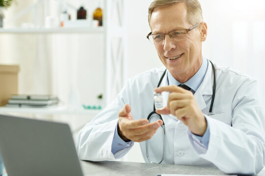 Joyful doctor holding covid-19 vaccine and using laptop in clinic - Powered by Adobe