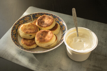A plate with cheese pancakes next to sour cream  on a marble mat on a black wooden table. A breakfast on a sunny morning.