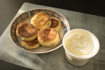 A plate with cheese pancakes next to sour cream  on a marble mat on a black wooden table. A breakfast on a sunny morning.