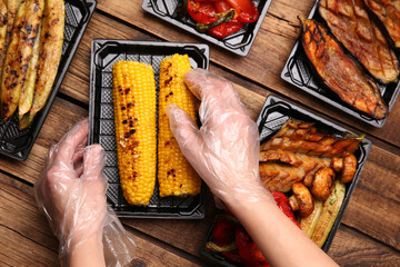 Waiter in gloves putting grilled corn cobs into containers at wooden table, top view. Food delivery...