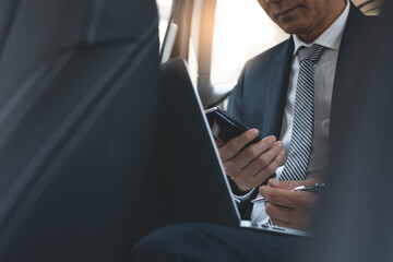 Businessman in black suit using mobile smartphone and working on laptop computer inside a car