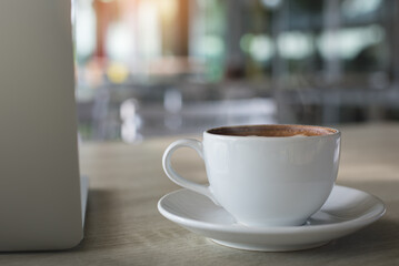 White cup of coffee and laptop computer on wooden table in coffee shop co-working space with nobody