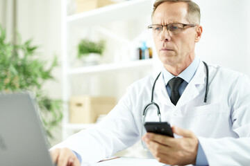 Serious doctor holding cellphone and using laptop at work