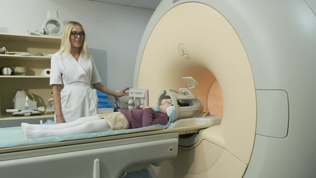 Woman doctor radiologist makes an MRI scanning of brain, head, neck for patient little girl, girl lies on automatic table, using modern equipment, coil on the head, headphones