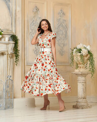 Young beautiful pretty elegant woman with long brunette hair wearing colorful dress with floral print standing and posing in bright interior