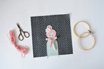 Handmade fabric applique with cute doll and bow, scissors, pink thread, embroidery hoop over white	