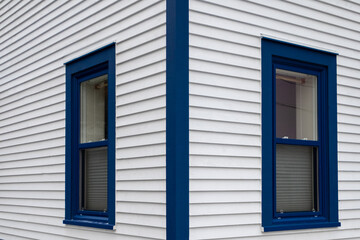 The exterior of a stark white cape cod clapboard horizontal wooden board siding wall with multiple blue trim double hung windows. The corner of the residential building sits on a concrete footing.