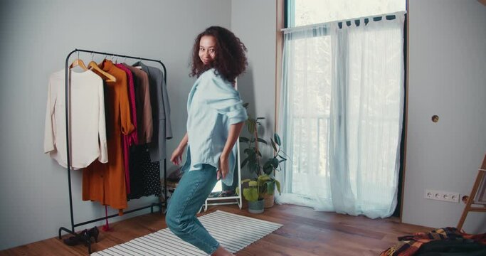 Beautiful young excited African American woman with curly hair dancing, looking at camera in cozy big wardrobe room.