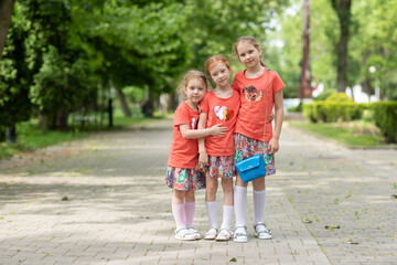 Three sisters stand in the center of the park hugging and saying something to each other in the ear, it's green on the street, the girls are dressed in bright orange dresses