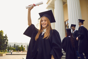 Happy smiling graduate girl dressed in black gown raises up her hand holding a diploma. Student...