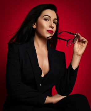 Portrait sexy adult brunette woman in official suit with deep neckline sitting holding her glasses looking flirty at camera over red background. Fashion, style, trendy business look for female concept