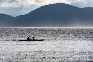Fototapeta premium Two people canoeing in the late afternoon with a fog in the bay of the city of Santos. In the background, the mountains and blue sky.