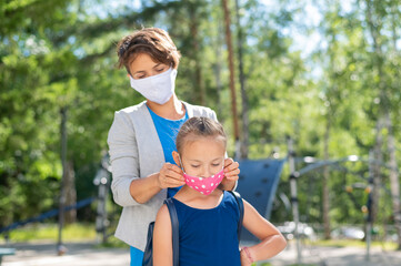 Caucasian woman puts a protective mask on her daughter outdoors. Caring mother helps to wear a mask schoolgirl near the children's playground. Quarantine during coronavirus.