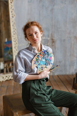 Fototapeta premium Portrait of a red-haired professional artist with a brush and palette, writing on canvas in the studio. A female artist draws in the workplace.