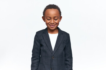 Happy childhood, joy and positive emotions concept. Isolated image of adorable cheerful black little boy in formal clothing looking down with shy smile. Timid dark skinned child smiling in studio