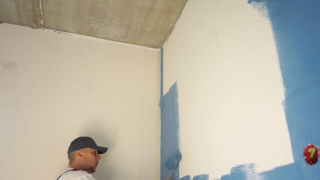 Male painter painting a white wall with a roller in blue color renovations or construction concept.